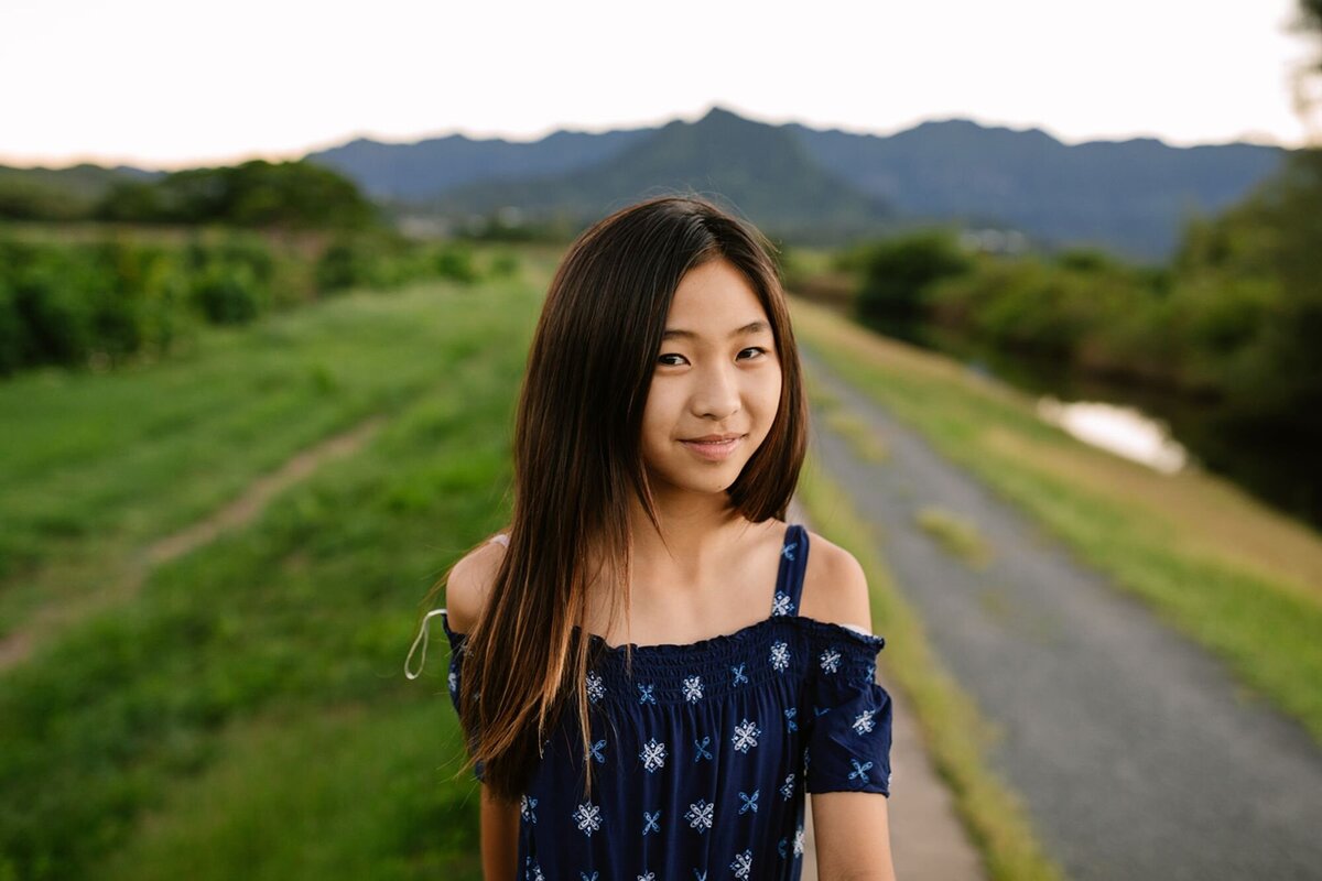 A girl walks along a road with mountains and green lush, tropical forest around her.