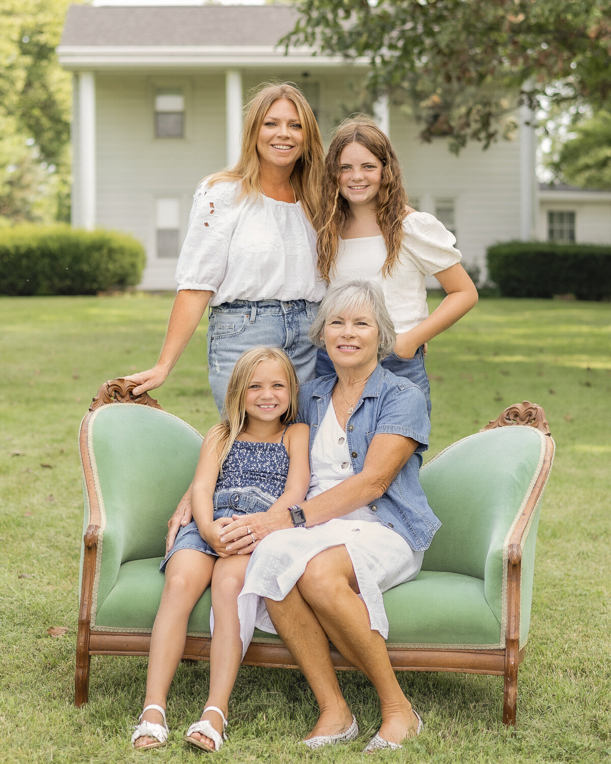 Mom and oldest daughter are standing behind the vintage green couch, grandma and youngest granddaughter are sitting in the vintage green couch in the grass with their family home in the background, everyone smiling at the camera.  Daughters are sitting on the vintage green couch.  Everyone is looking at the camera smiling.  Mom is wearing a jean skirt, white half sleeve shirt, and has long blonde hair.  Older daughter is wearing blue jean shorts, a white short sleeve shirt, and has long brown hair.  Grandma is wearing a white button up dress with a jean jacket over it, with short white hair.  Youngest daughter has a dark blue and white tank top style shirt with a blue jean skirt, and has medium length blonde hair.