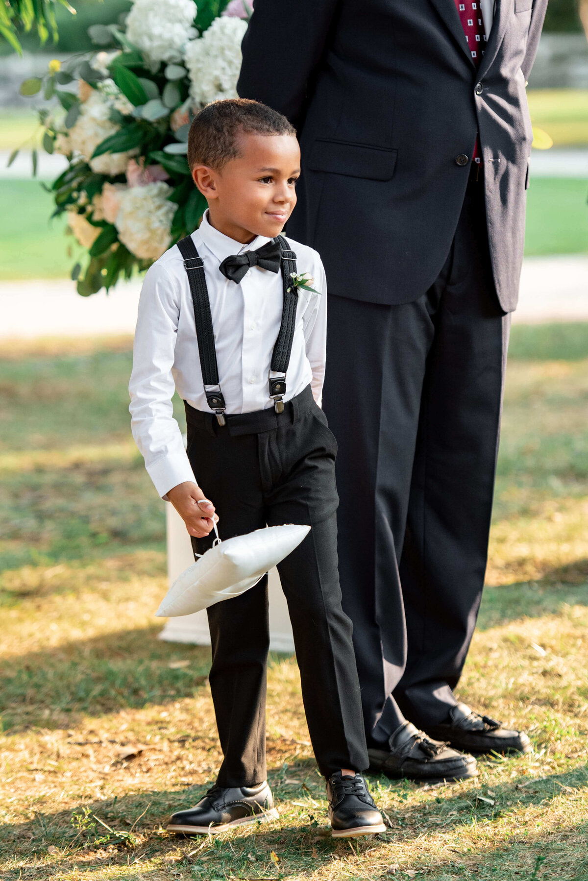 A young boy dressed in a white shirt, black suspenders, and bow tie holds a white pillow, standing outdoors next to an adult in a suit. A floral arrangement is visible in the background.