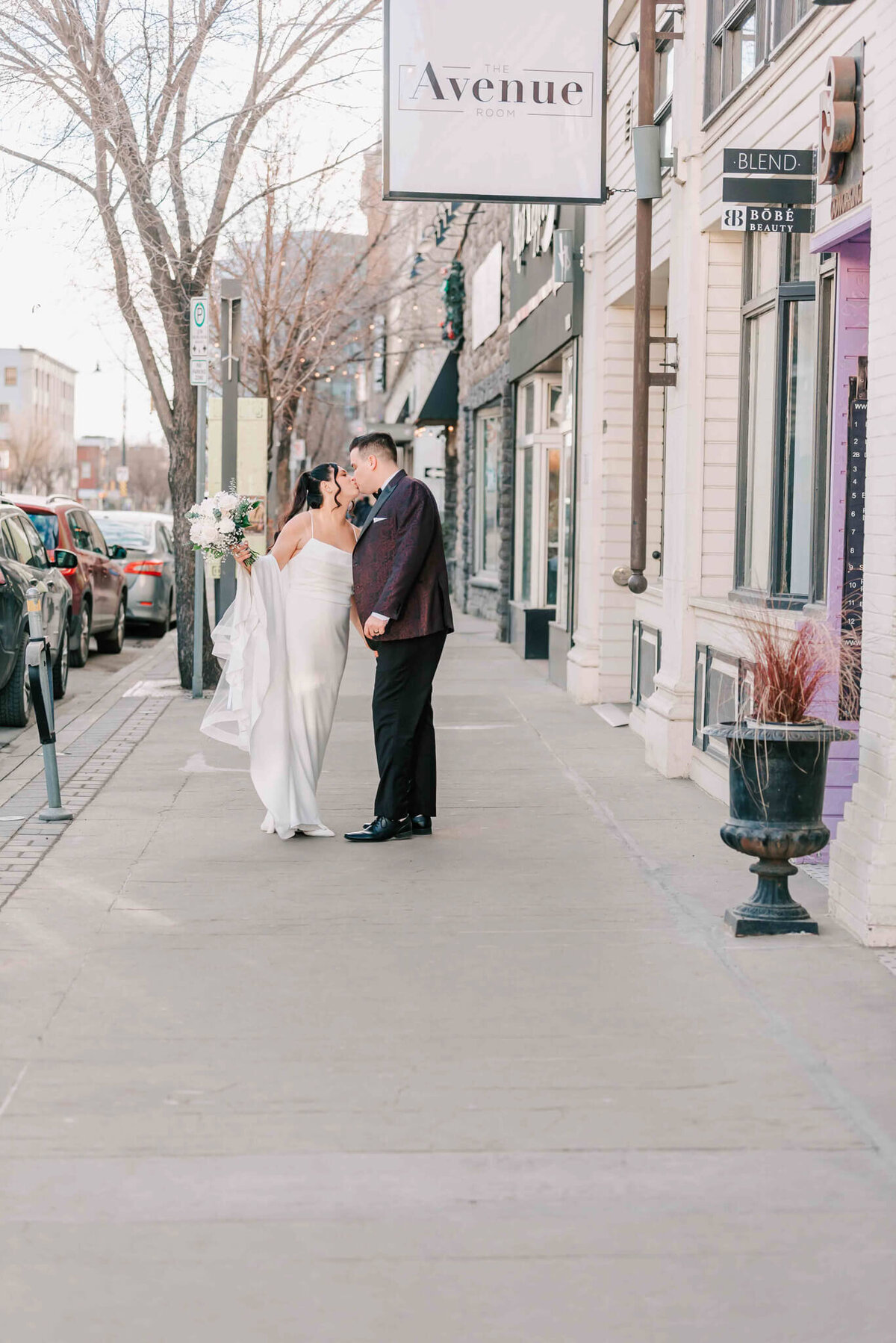 Bride and groom share a kiss standing on the sidewalk downtown. 