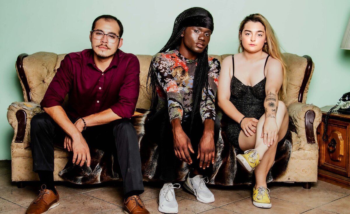 Musical trio portrait The Kidz sitting tightly together against tan sofa pale green wall background