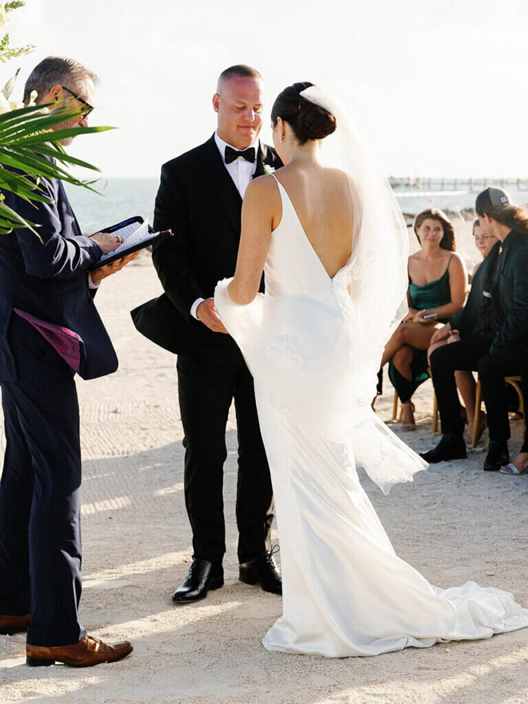 A romantic moment as the bride and groom hold hands while exchanging vows at their elegant beachfront ceremony, with the ocean waves in the background. Destination wedding photography by Claudia Amalia Photography in the Florida Keys.