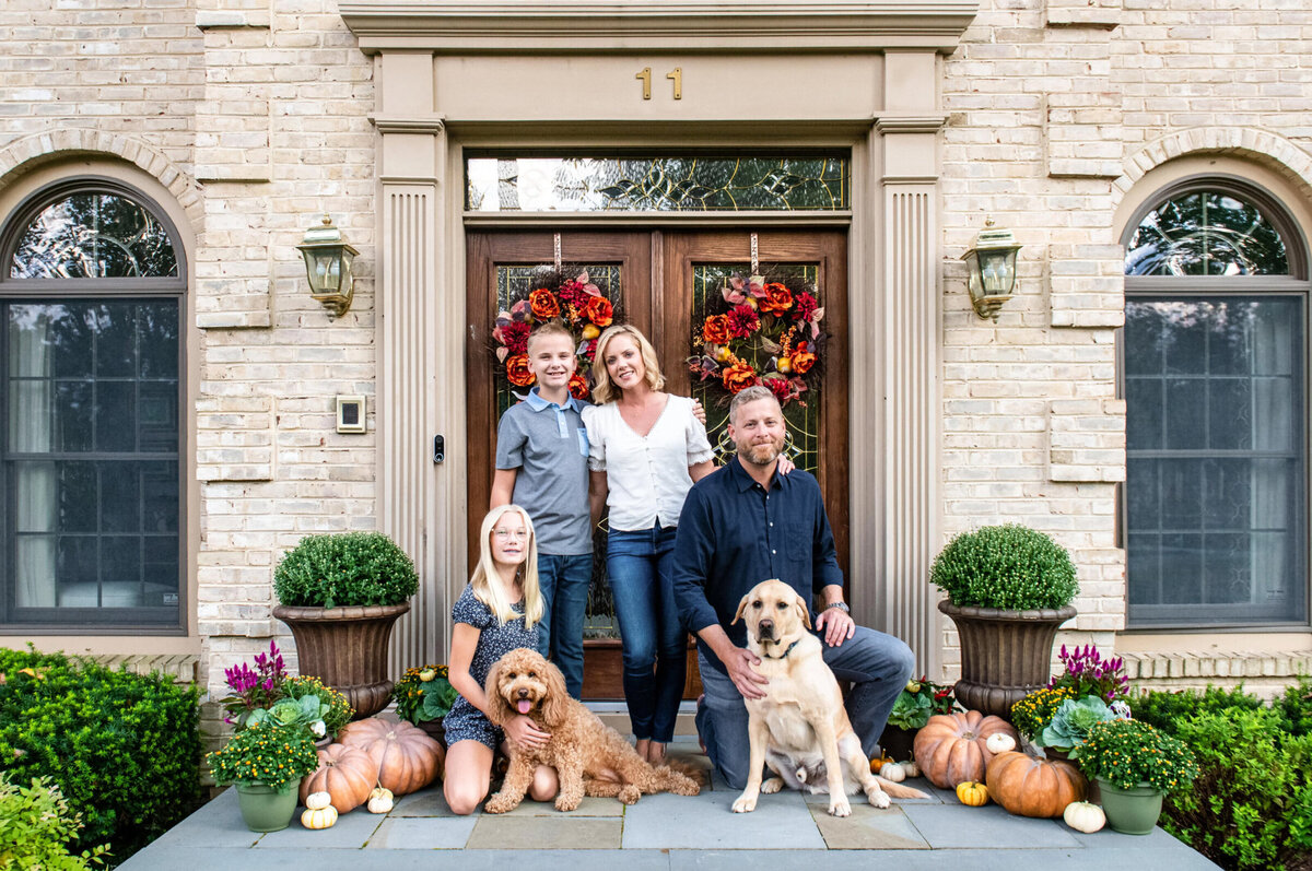 Family of four posing with their two dogs in front of their home that is decorated for fall