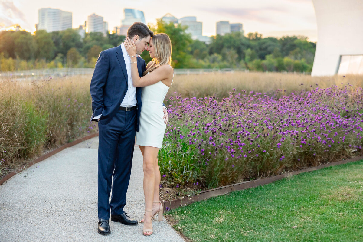A couple stands on a pathway beside a field of purple flowers. The man, in a dark suit, and the woman, in a white dress, lean toward each other with affectionate smiles. The backdrop features a city skyline and greenery under a soft sky.