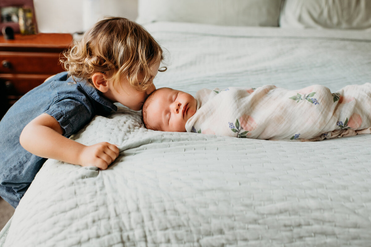Newborn Photography, Baby is laying on the bed, big brother is standing up and kissing baby on the head.