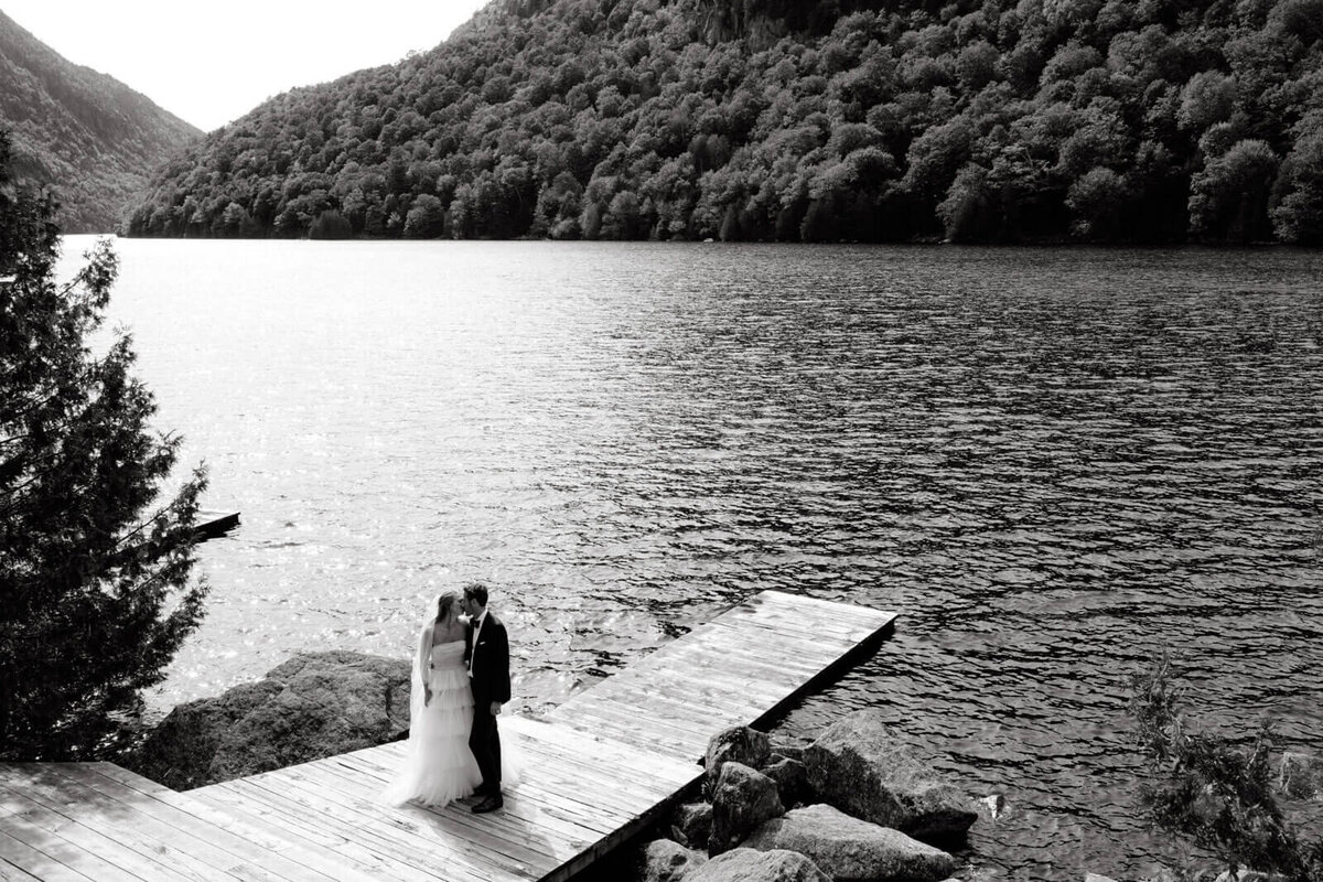 Black and white photo of the bride and groom kissing on a wooden dock overlooking the waters and mountains at The Ausable Club.