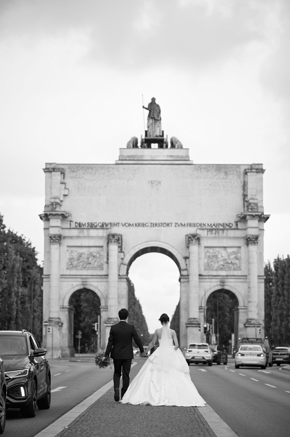 Ein japanisches Hochzeitspaar geht Hand in Hand auf das Siegestor in München zu