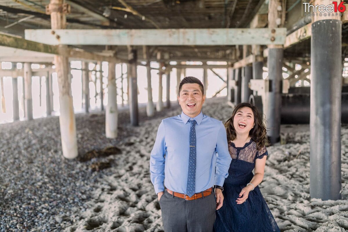 Engaged couple breaks out in laughter while posing under the San Clemente Pier