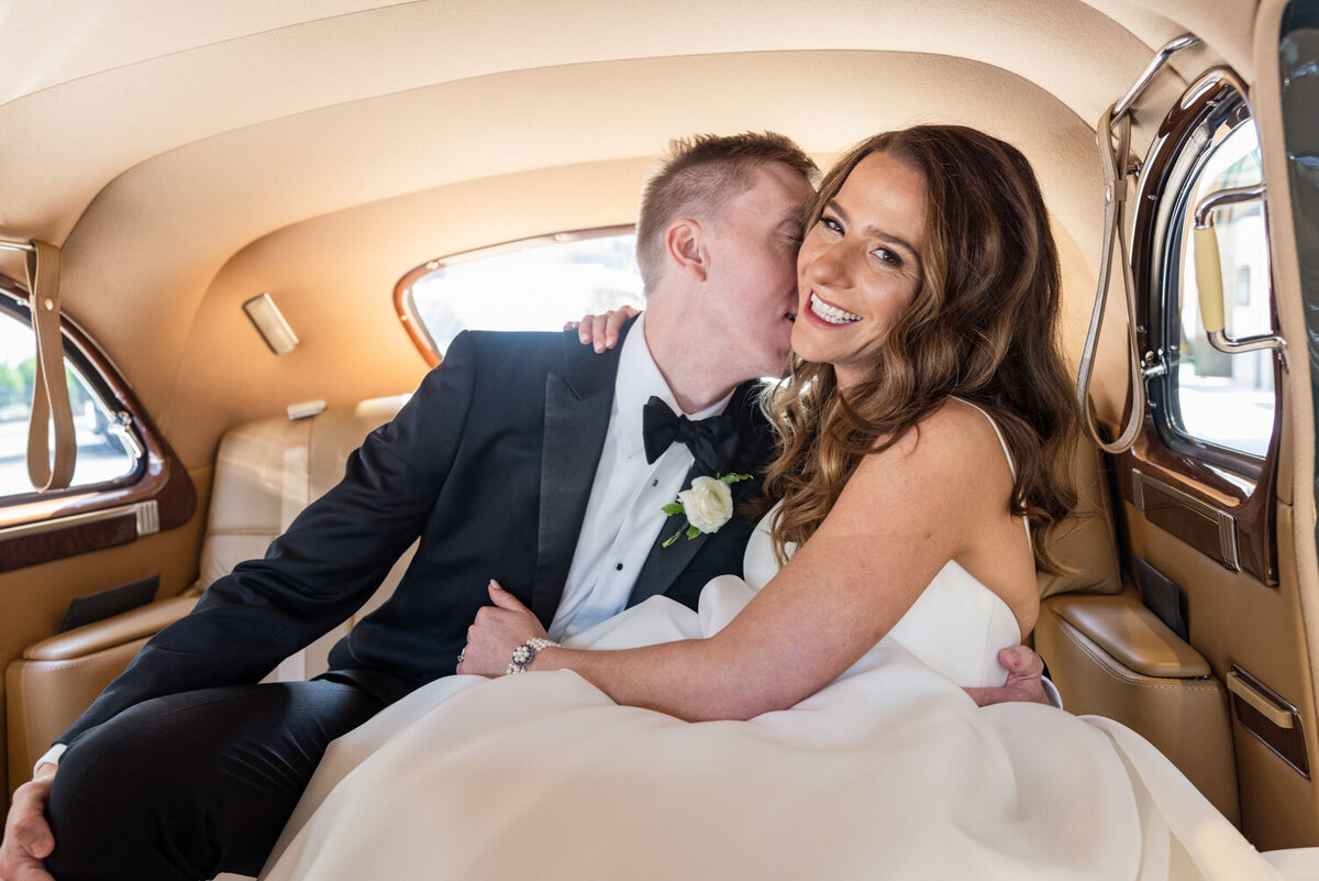 A bride and groom are sitting in a car and the bride is kissing the groom on the cheek.