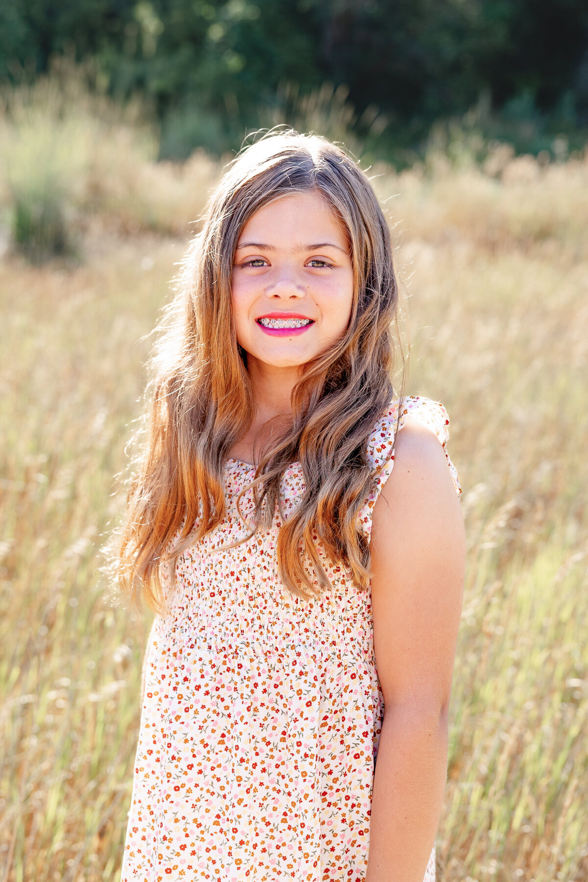 A young girl with long, wavy brown hair beams at the camera, wearing a floral summer dress. The golden backdrop of the field enhances her radiant smile.