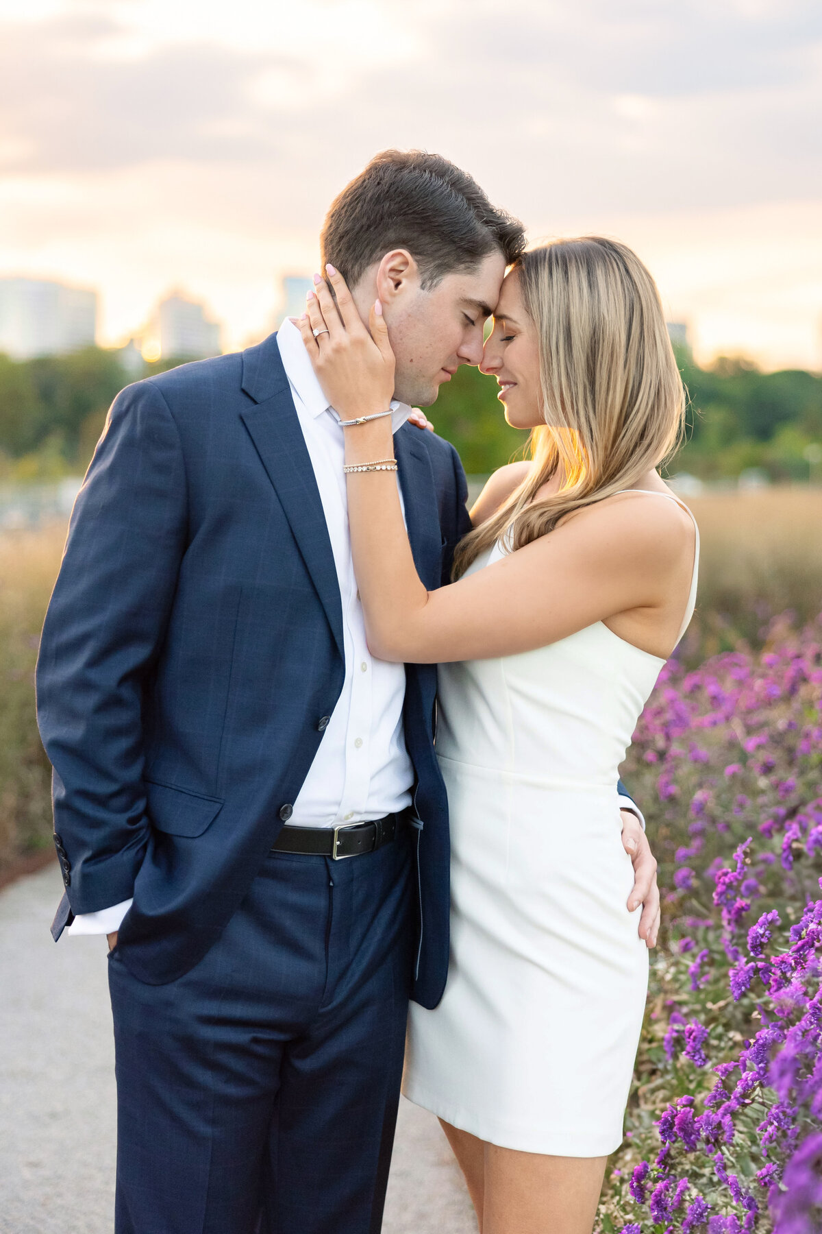 A couple stands on a garden path, embracing and leaning in for a kiss. The man is wearing a navy suit, and the woman is in a white dress. Purple flowers line the path, with a cityscape in the background under a soft, cloudy sky.