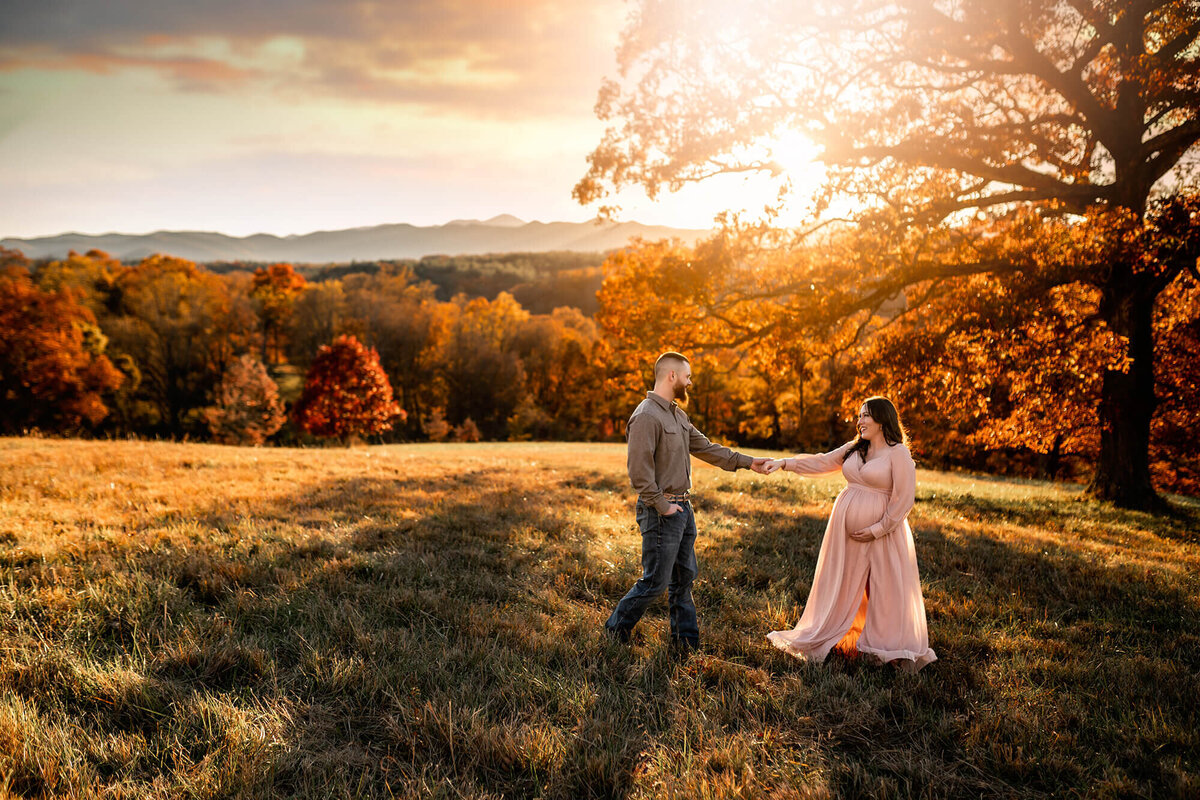 A pregnant woman in a pink dress dancing with her husband in the setting sunlight