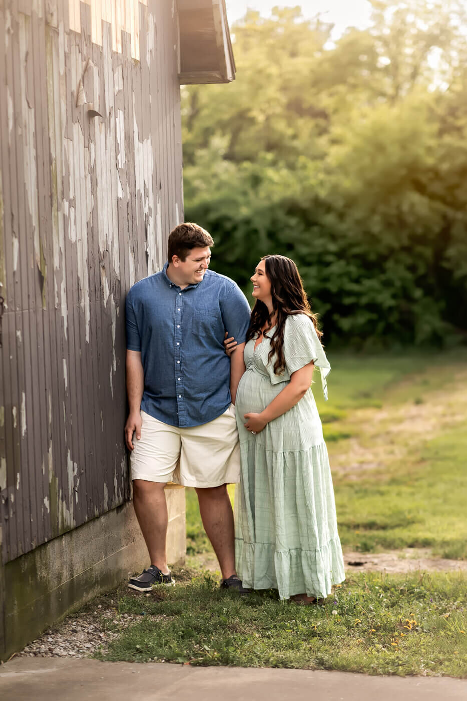 A family of four smiles together in a studio, perfect for maternity portraits. The father holds a young boy on his shoulders, while the mother—captured by a Bloomington maternity photographer—holds another young boy; all wear white tops and jeans.