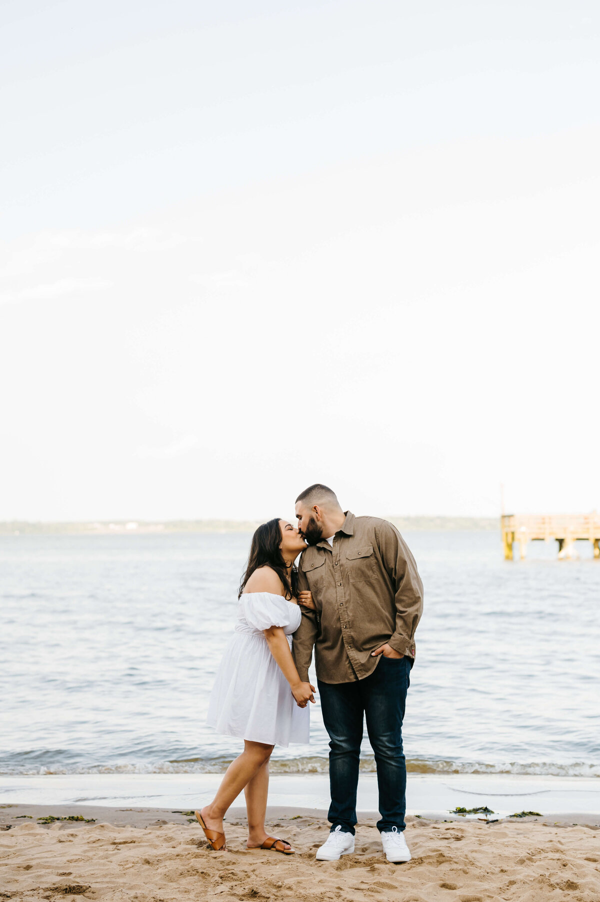 woodbridge lake beach engagement photos with man and woman holding hands while standing side by side and the woman grabbing the mans arm with her other hand and leans into him to kiss