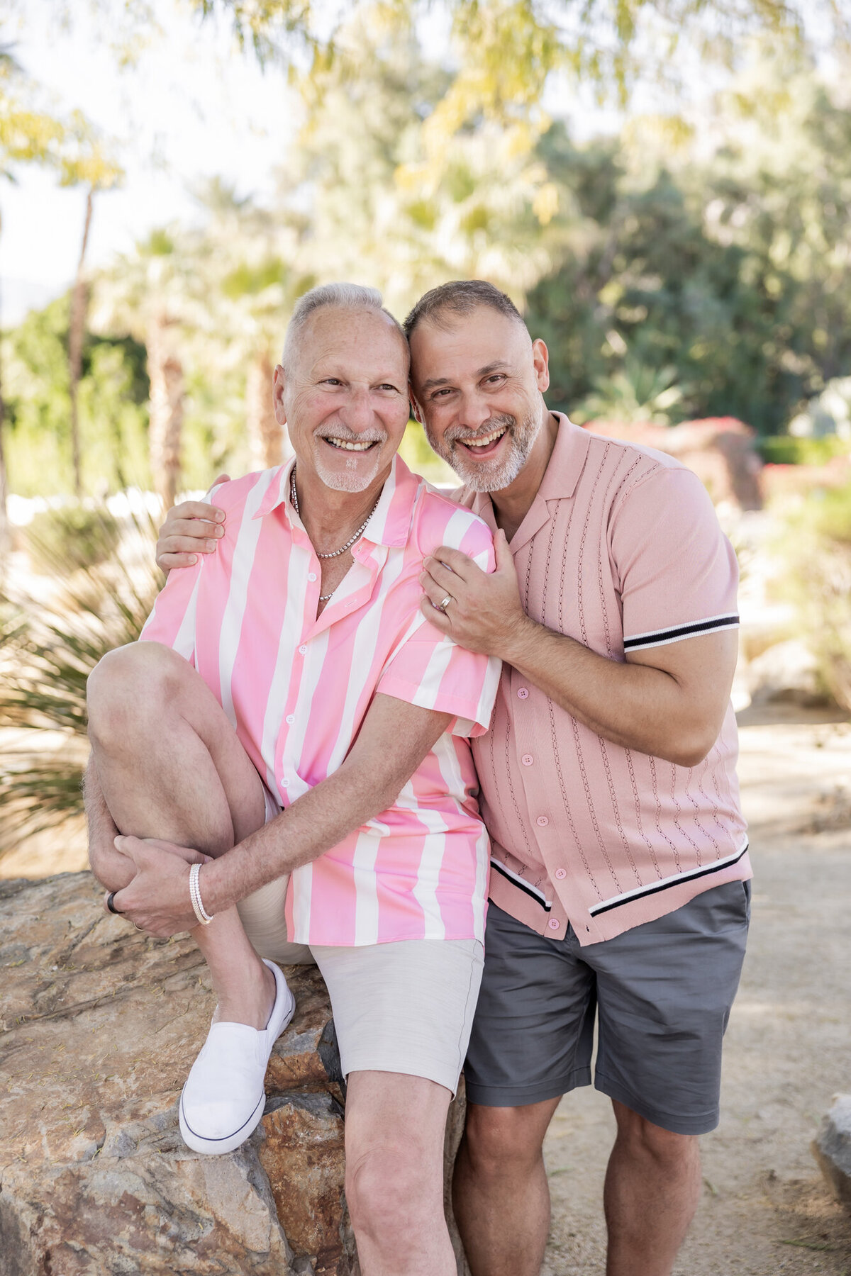 LGBTQ+ couple holding hands and laughing in soft desert light, dressed in coordinating pink and tan tones.
