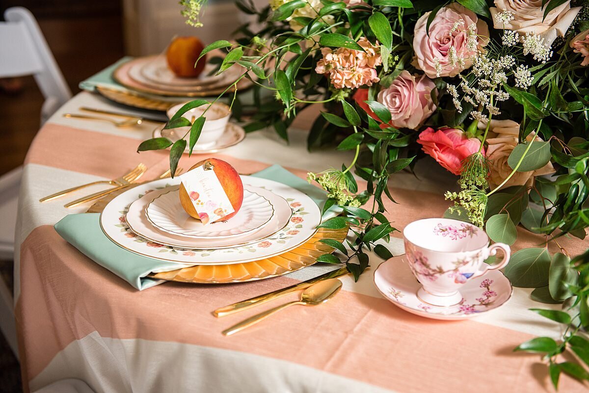 Peach and white striped table cloth topped with a gold charger, mismatched vintage china dinner plate, mismatched vintage china salad plate, mismatched vintage dessert plate and a mint green napkin. On top of the plates is a fresh peach with a seating card written in hand drawn calligraphy. The table is also set with matte gold flatware and a mismatched china tea cup.  There is a large floral centerpiece filled with lush greenery, peach, blush, pink, ivory and white flowers.