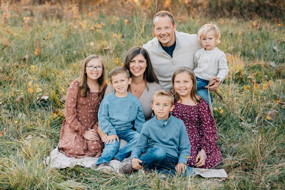 Family of 7 sitting together outdoors, surrounded by colorful fall foliage.
