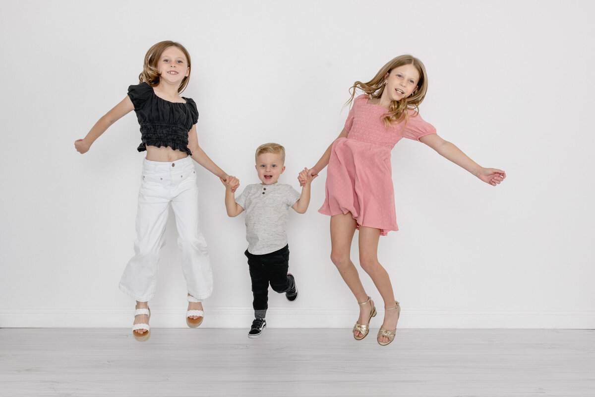 Three young siblings with blonde hair holding hands and jumping off of the ground while smiling at the camera in a white studio in Tustin.