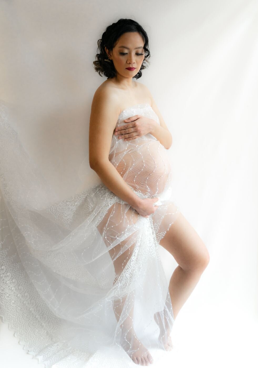 Pregnant woman wrapped in flowing white lace standing barefoot and cradling her belly with one hand while softly looking down