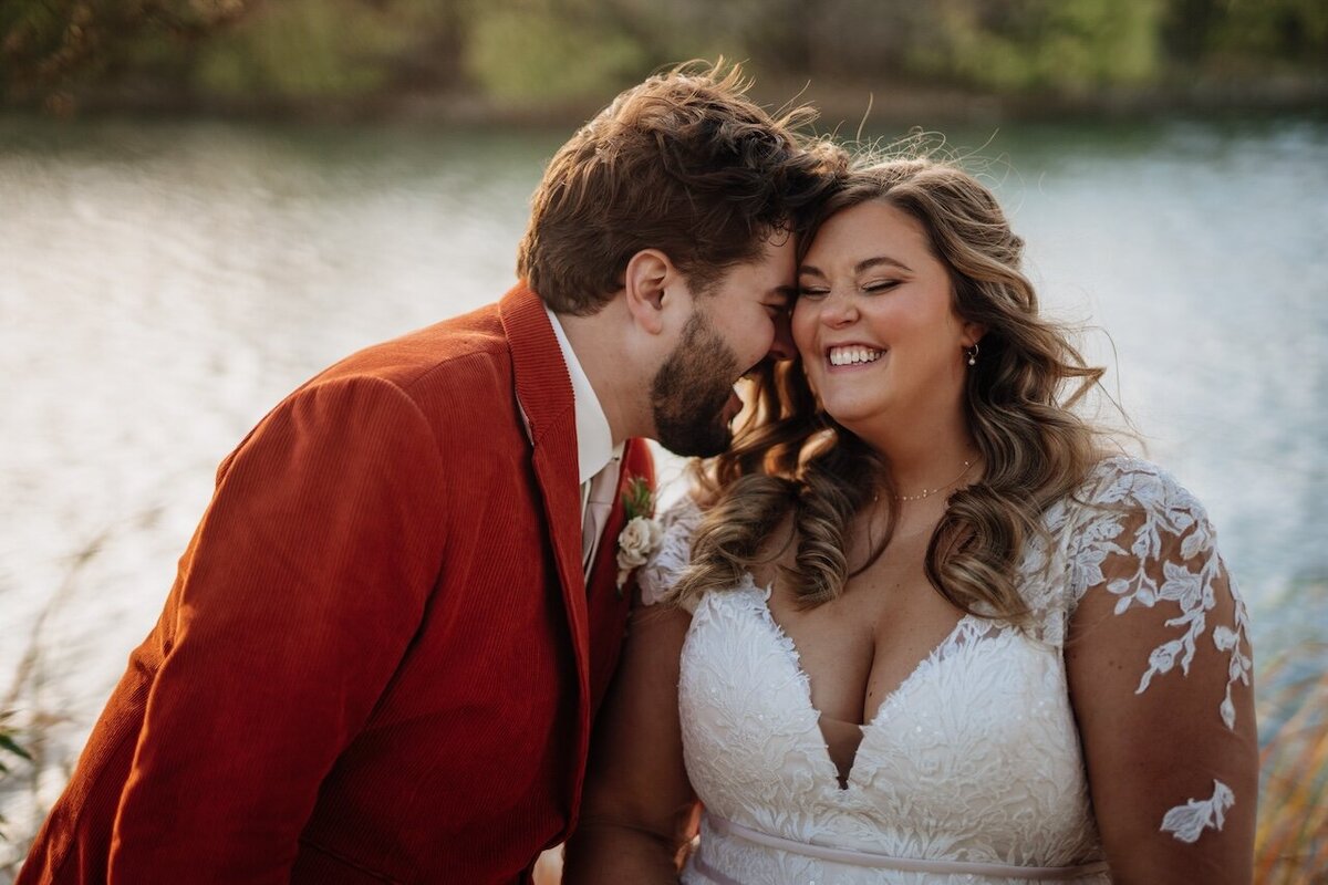 A bride and groom share a tender moment by a tranquil lake at sunset. The groom, in a striking rust-colored jacket and white shirt, leans in to kiss the bride, who beams joyfully. She wears a white lace gown with floral details, complementing her flowing, curled hairstyle. The golden hour light softly highlights their joyful expressions and the serene water backdrop.