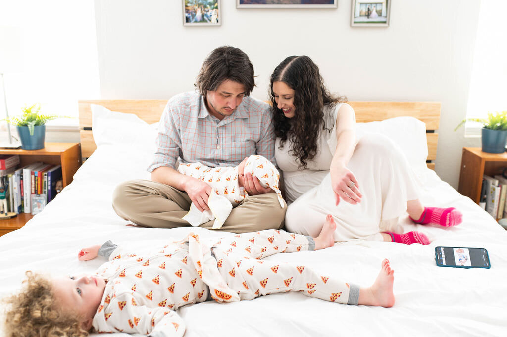 Parents sitting on a bed holding a newborn with a small child laying in front of them.