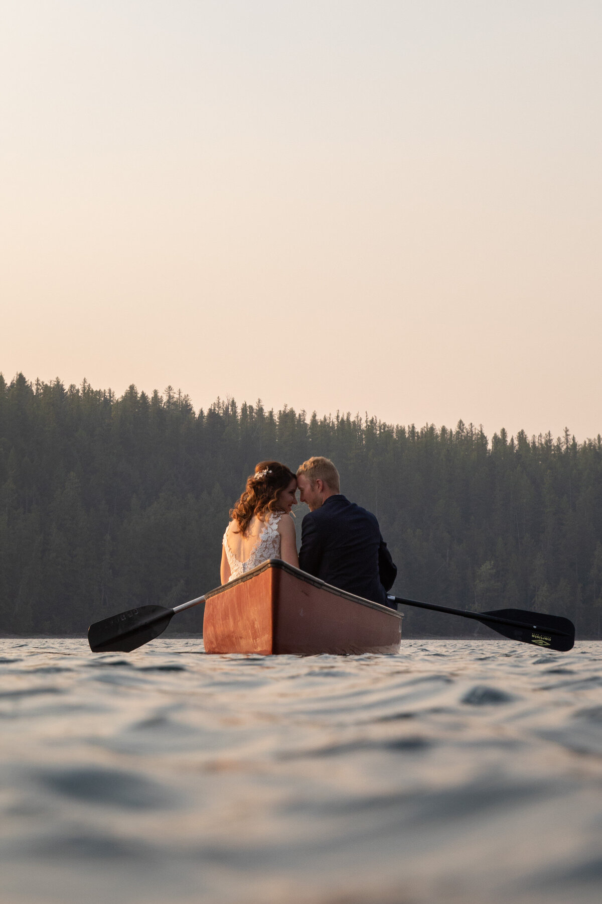 A bride and groom sit next to each other in a canoe with their foreheads touching.
