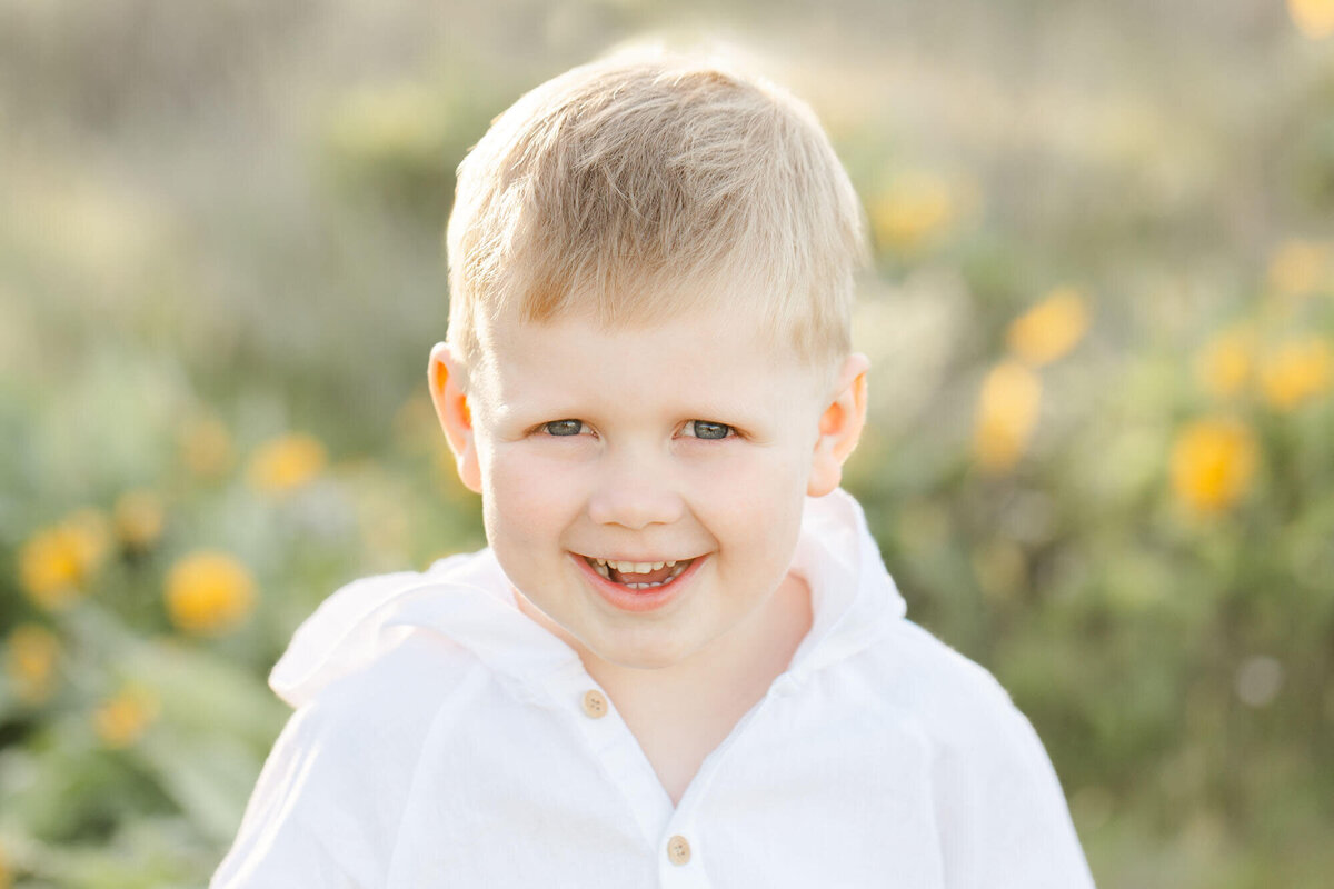 Four Year old boy dressed in white and sitting in a field of green grass and yellow wildflowers. This image is a close-up of his face and shoulders and the wildflowers are blurred out in the background. He has a big smile on his face and the sunlight is highlighting the rim of his head.