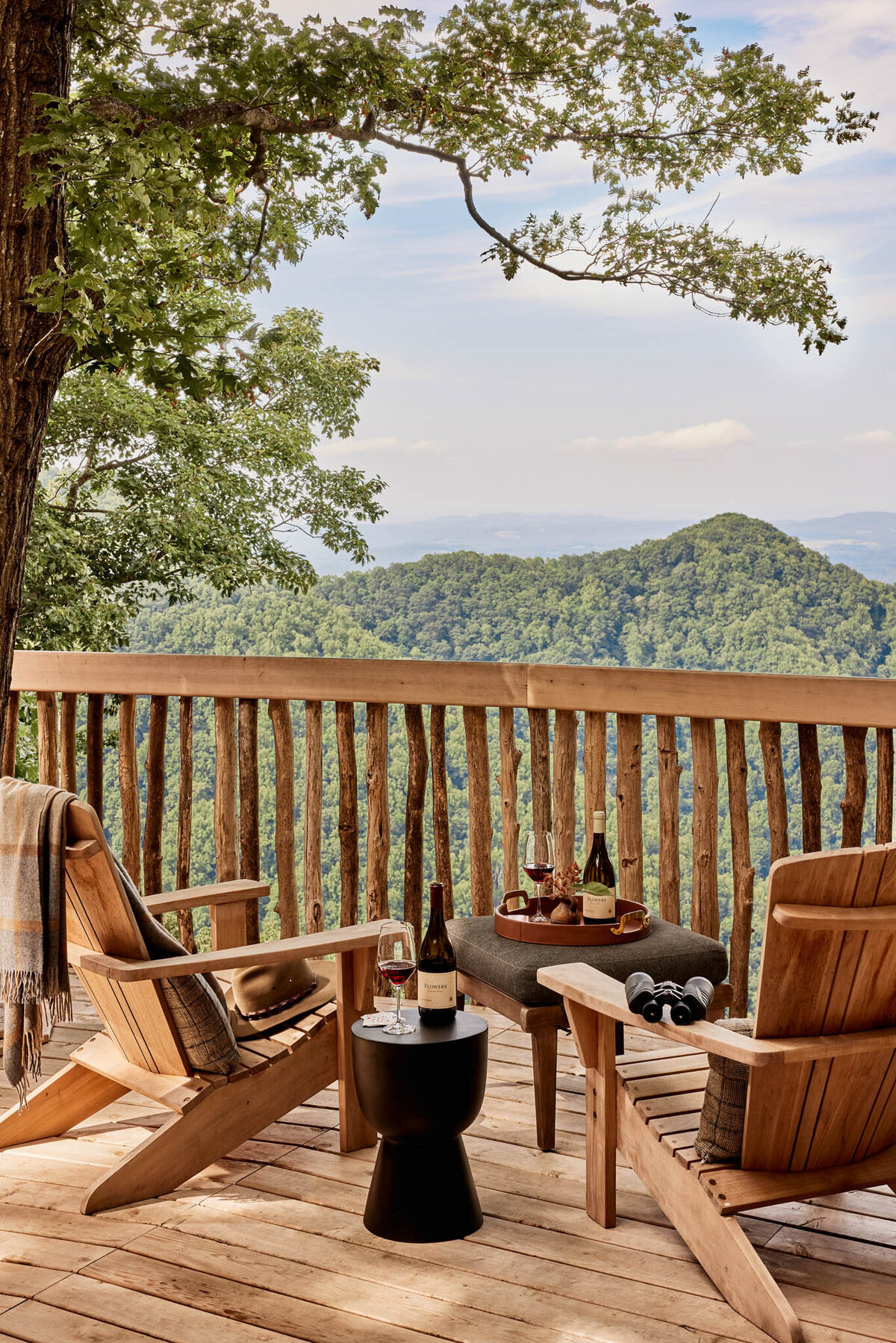 Rustic wooden deck overlooking lush green mountains. Two wooden chairs face outward, with a small black table between them holding wine bottles and glasses. A cozy blanket is draped over one chair, under a leafy tree canopy.