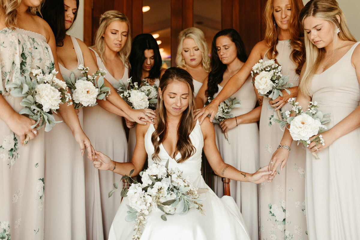 Bride sitting down and praying as she's holding hands with her bridesmaids as they surround her