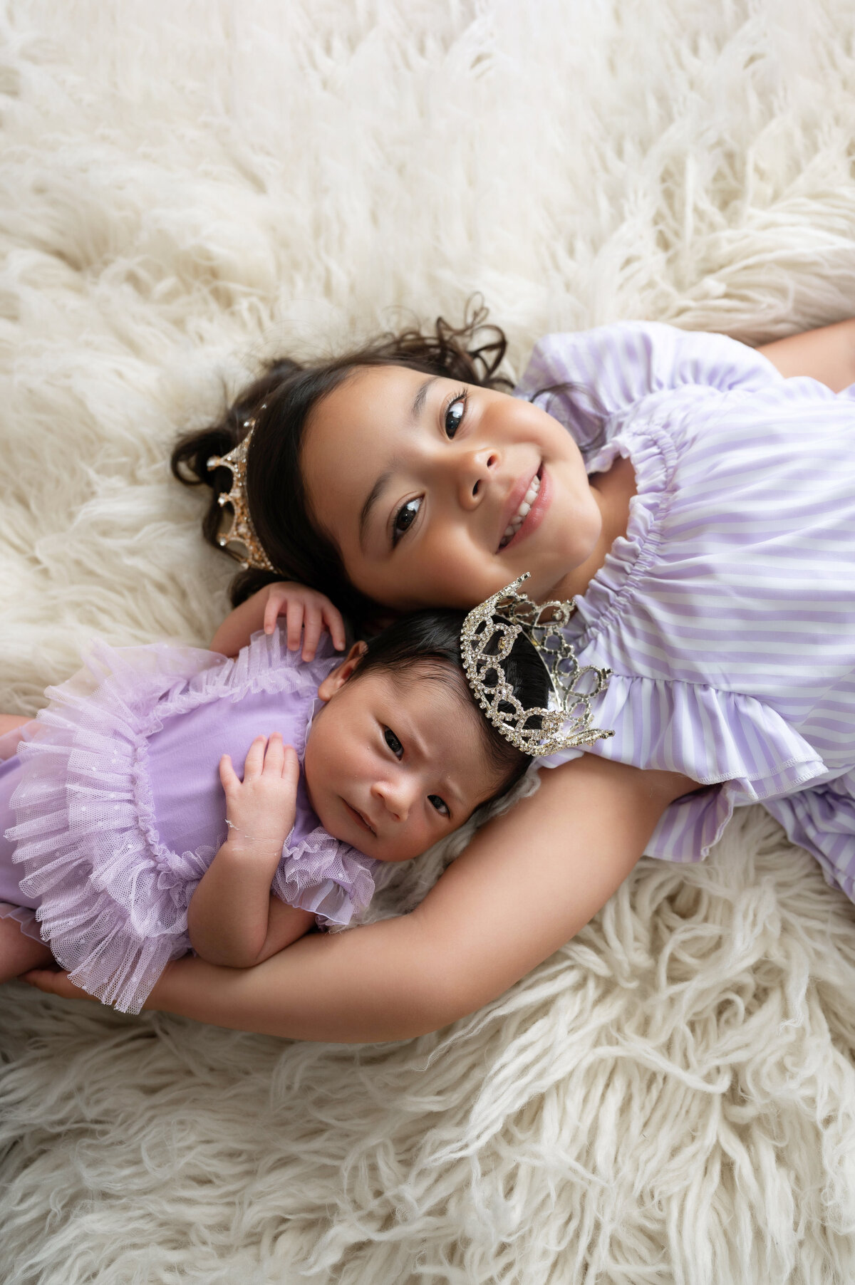 Toddler and newborn baby girl wearing matching lilac dresses and coordinating crowns, lying on an off-white rug.