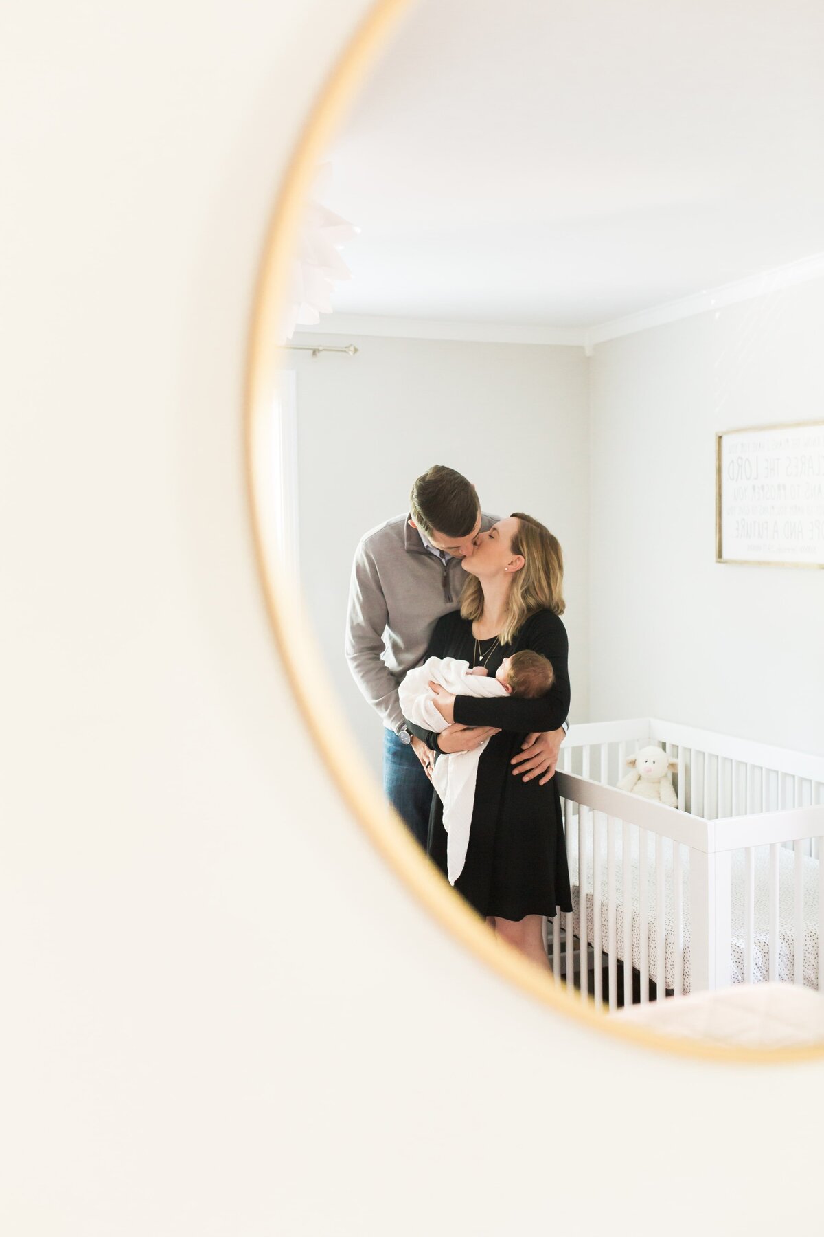 Two adults sharing a kiss while holding a baby in a nursery, as seen through a circular mirror, captured by an at-home newborn photography session.