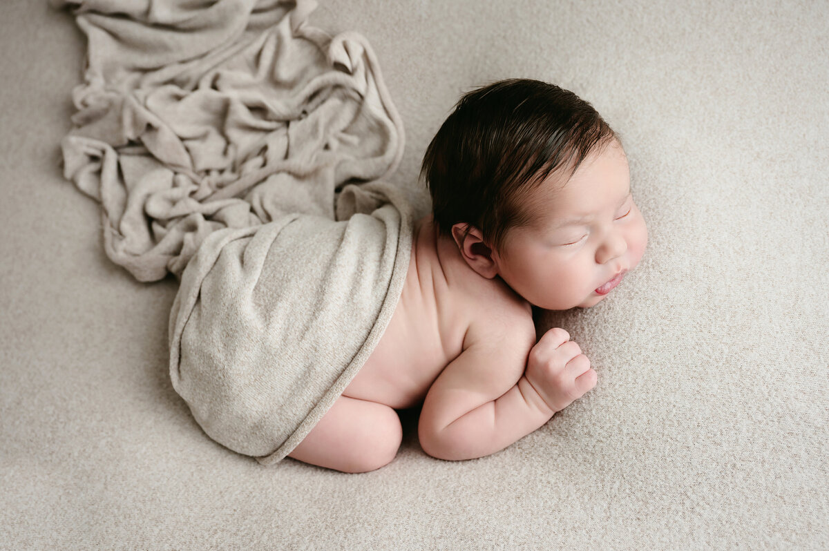 This adorable newborn baby boy photography session in Bridgeville, PA features a serene moment with the baby wrapped in a soft beige blanket. Captured in a cozy setting, this image showcases the peaceful beauty of a newborn’s first days.