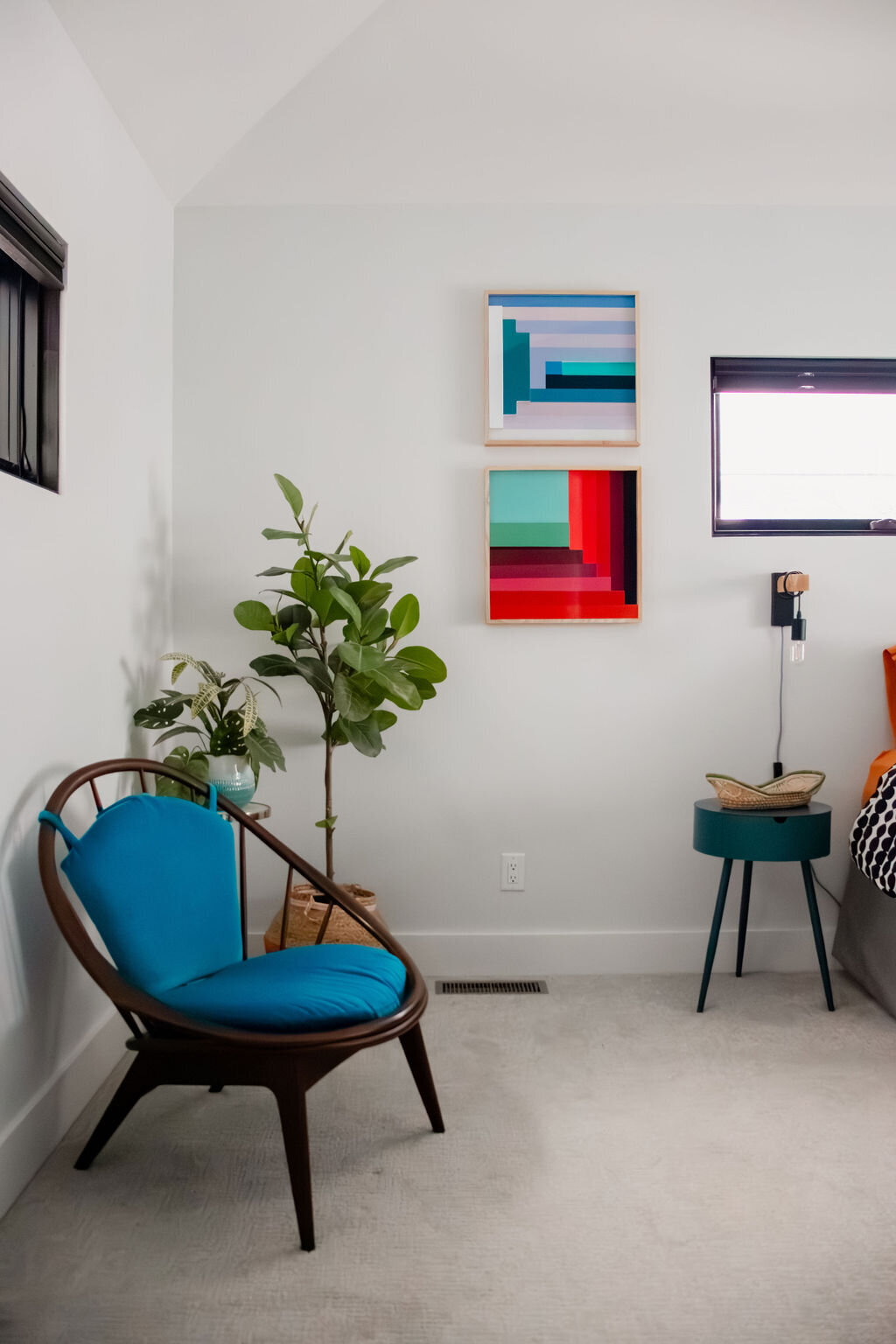 A bright, modern bedroom with white walls and a vaulted ceiling features a dark wood round-backed chair with a vibrant blue cushion. Next to the chair, a small glass-topped table holds two potted plants: a peperomia and a small fiddle leaf fig tree in a woven basket. On the wall above, two framed abstract artworks with geometric shapes in shades of blue, teal, red, and green are hung. To the right, a small round teal side table with black metal legs holds a woven basket. The edge of a bed with black and white patterned bedding and orange sheets is visible. A small black-framed window is above the bed, and another is to the left of the chair. The floor is covered in light gray carpet. A white electrical outlet is visible on the wall.