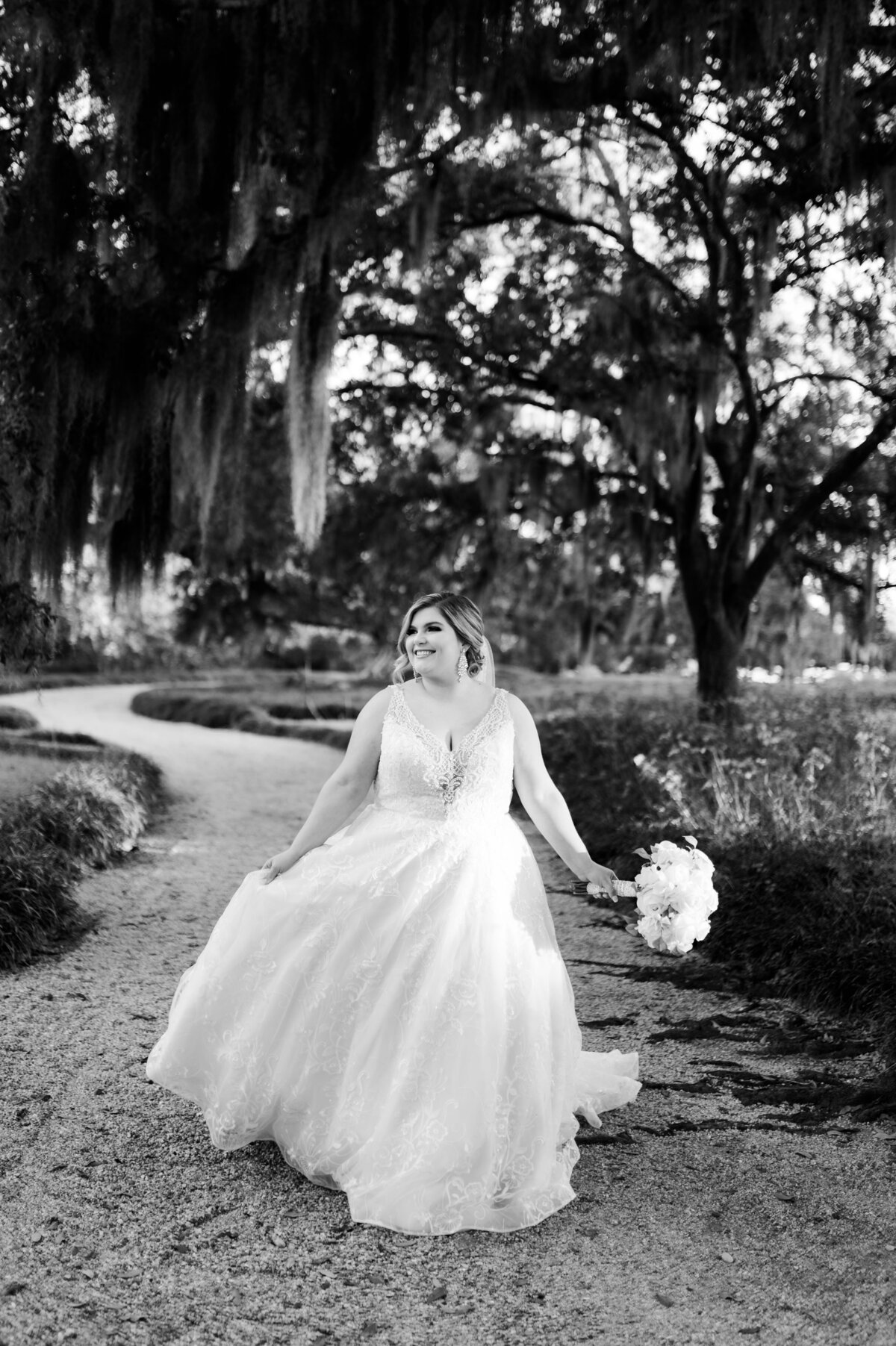 Little Rock wedding photographer captured bride walking down a path while holding her wedding dress with one hand and her bouquet with the other as the moss on the trees drapes down over her