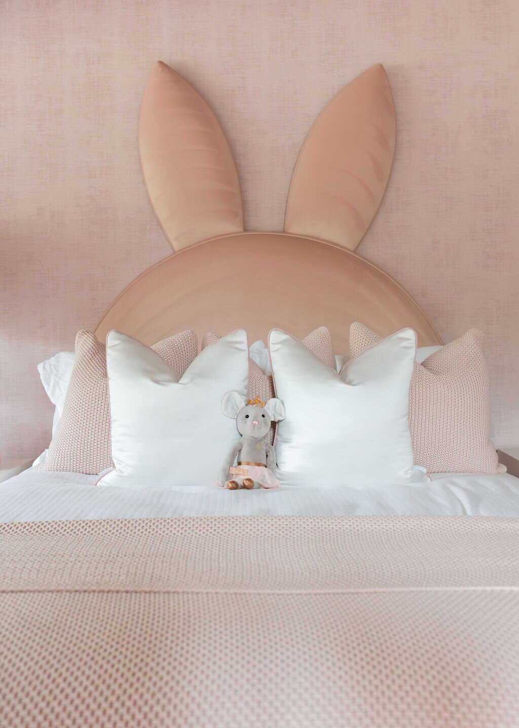 A cozy girl’s bedroom features a soft pink color palette, with a bed adorned with a unique headboard shaped like bunny ears.