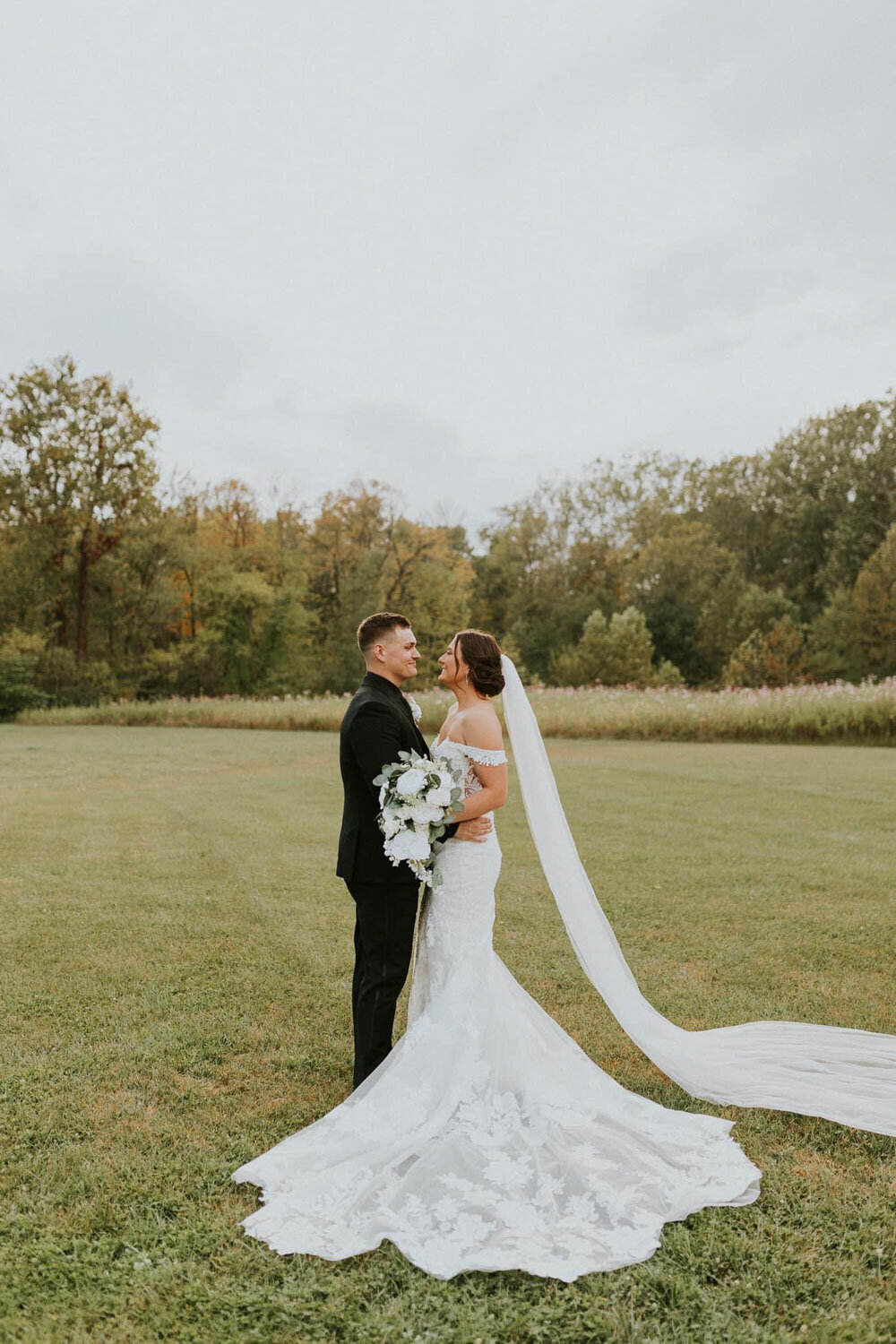 Bride and groom standing together in a green field with a long veil