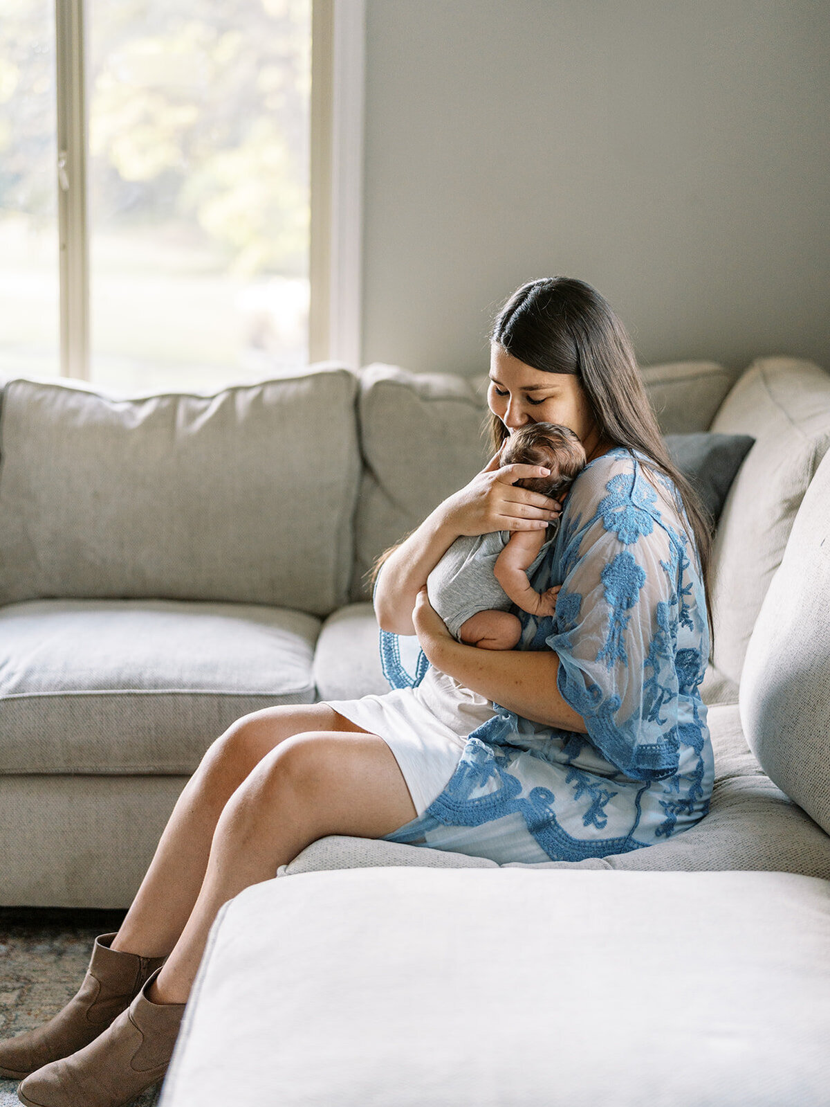 A woman sits on a couch at her home holding a baby up to her shoulder.