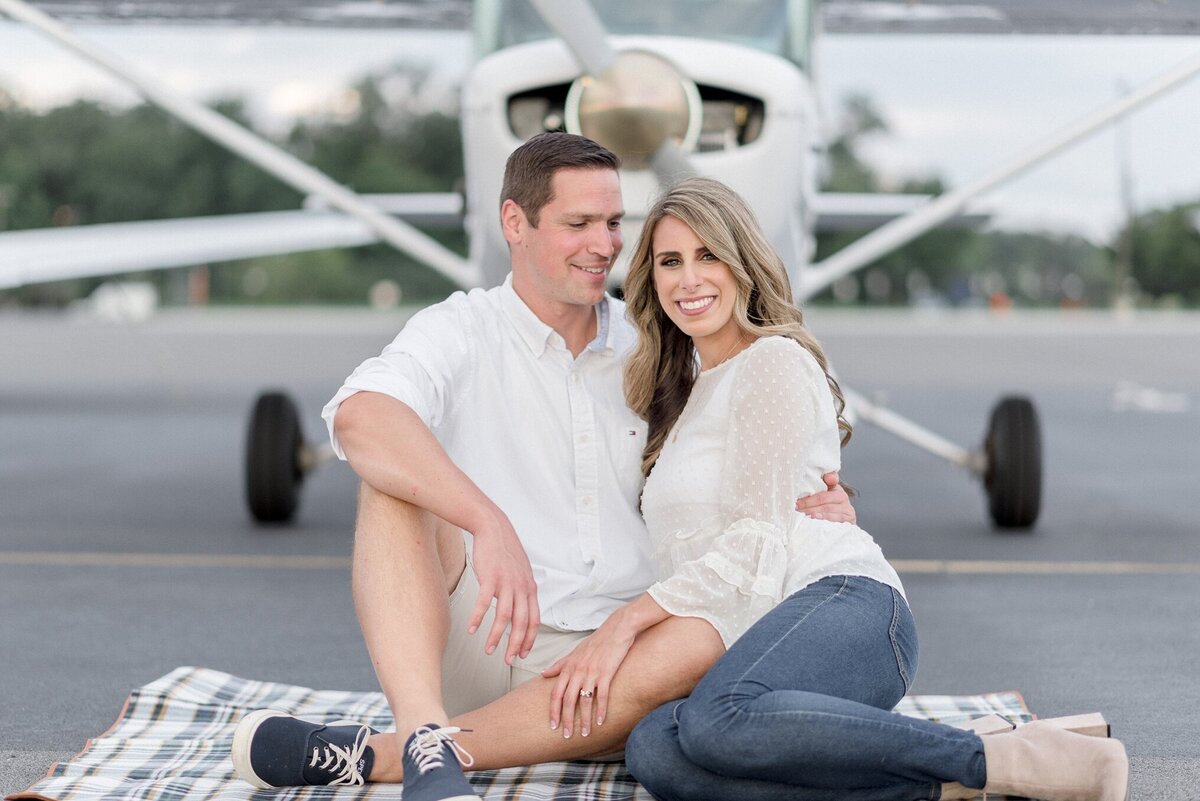 Engaged couple sitting & cuddling in front of air plane.