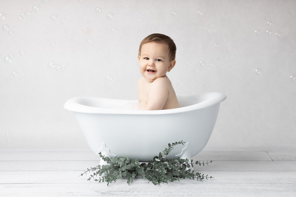 Baby boy enjoys a bubbly bath in a white vintage tub with eucalyptus leaves at the base, surrounded by floating bubbles during a DFW milestone photography session.