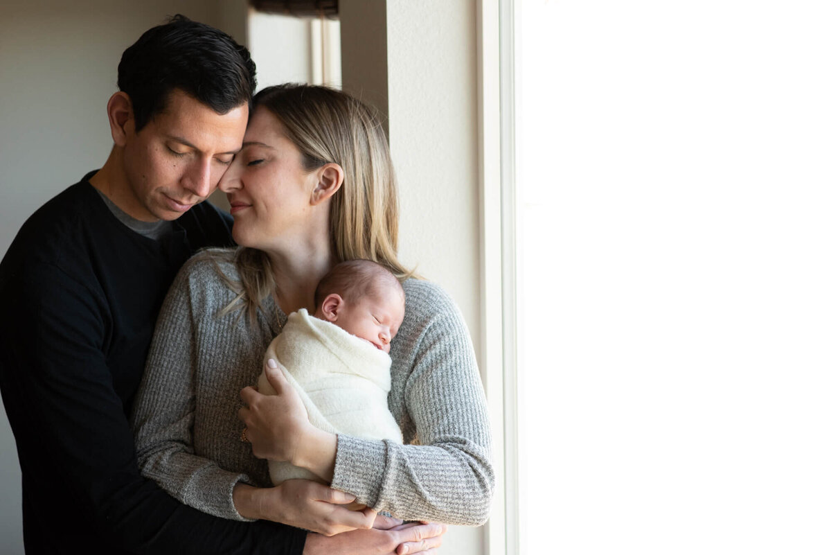 Happy new parents snuggle while standing in a window with their sleeping newborn baby