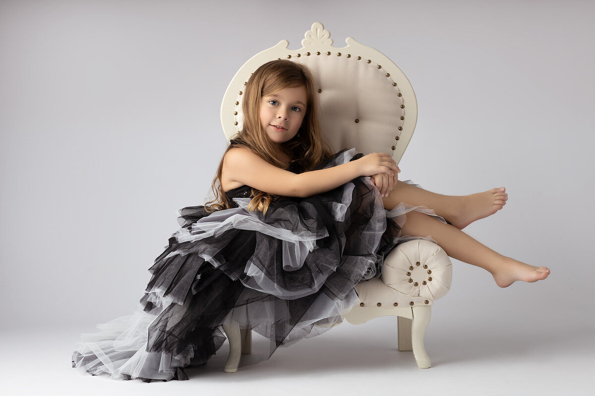 A young girl in a black and gray ruffled gown sitting on an elegant chair during her photo session.
