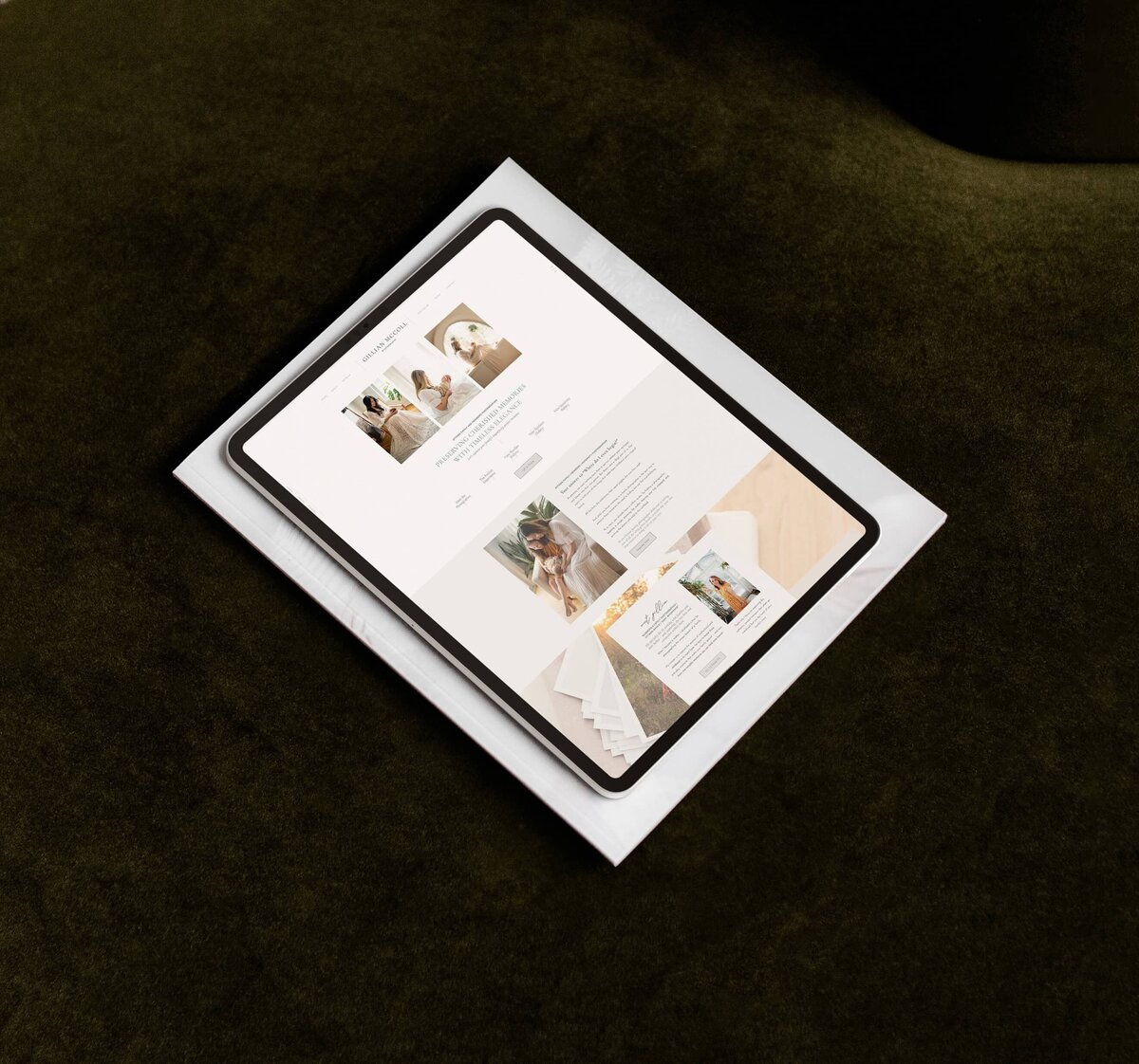 A tablet is placed on a stack of papers. The screen displays a webpage with images, text, and a light-colored layout, specializing in brand and identity services for luxury photographers. The background surface is a dark green fabric.