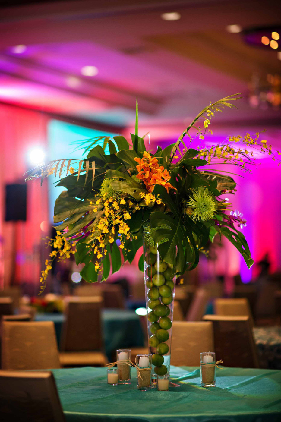 tall centerpiece with tropical leaves, purple orchids, in vase filled with limes on turquoise linen on turquoise linen