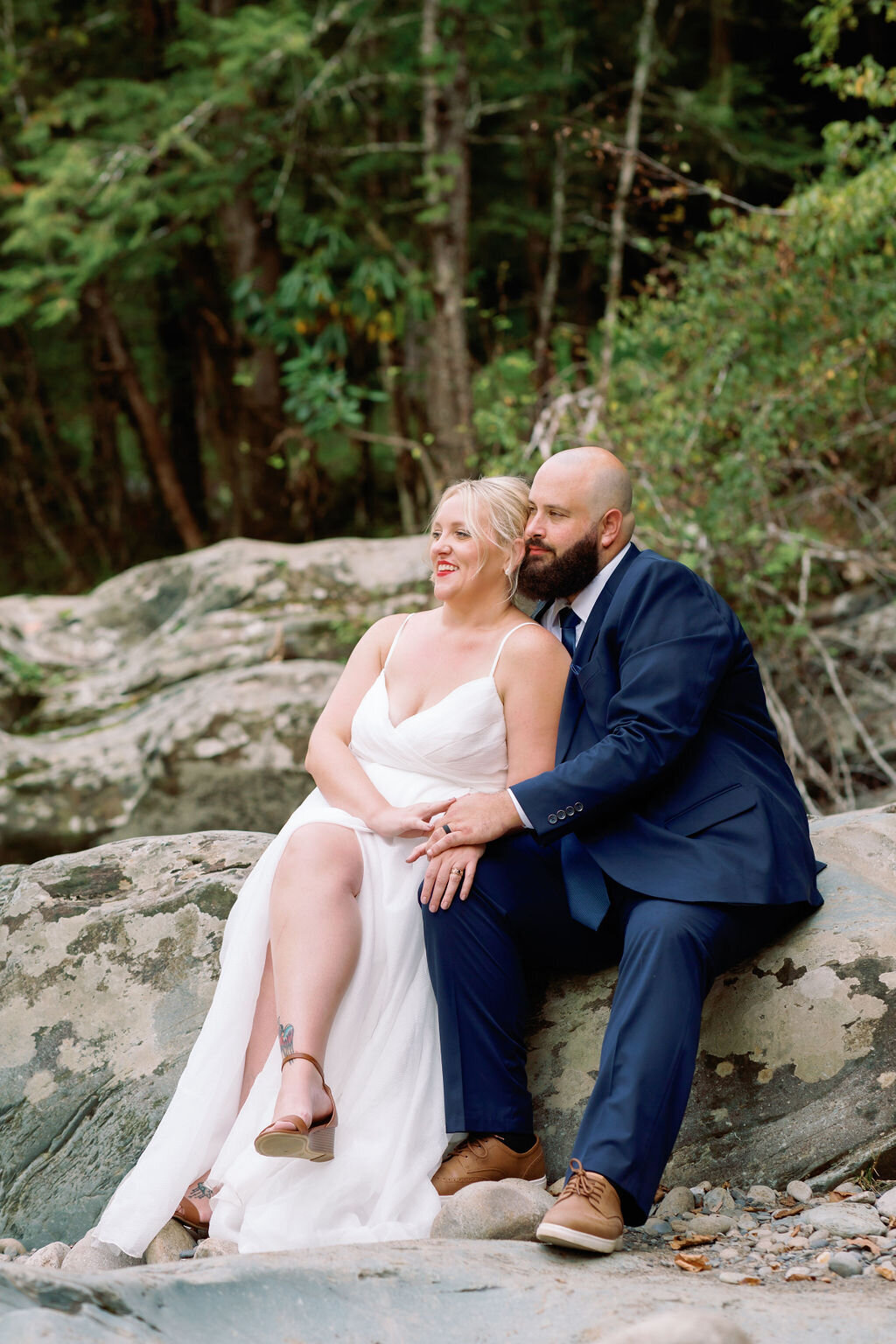 Bride and groom sitting close together on a large rock at Greenbrier, embracing and enjoying a quiet moment during their eloping to Gatlinburg celebration, with forest trees in the background.
