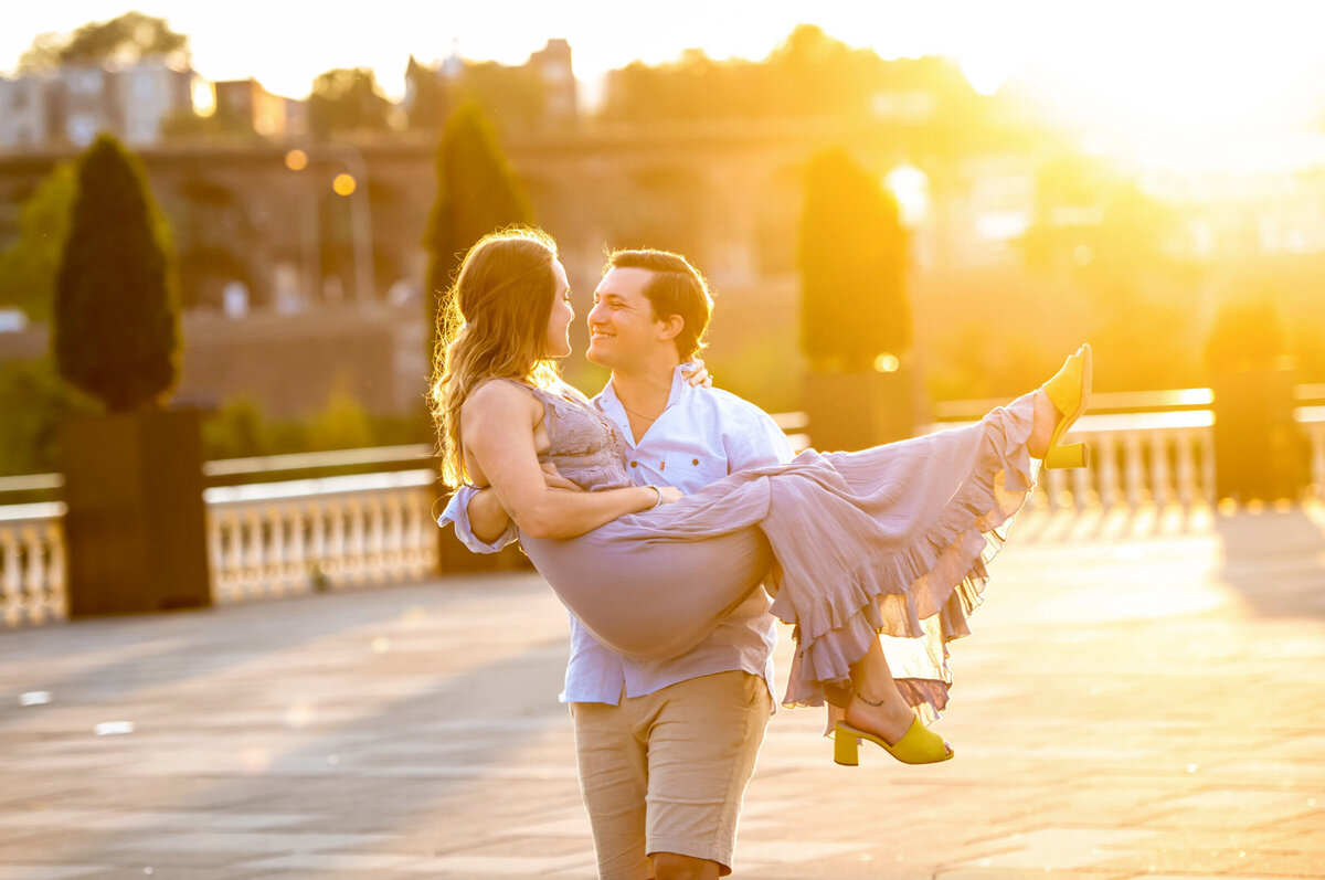 Man, carrying a woman in a purple dress with golden sunlight behind them and Fairmont waterworks