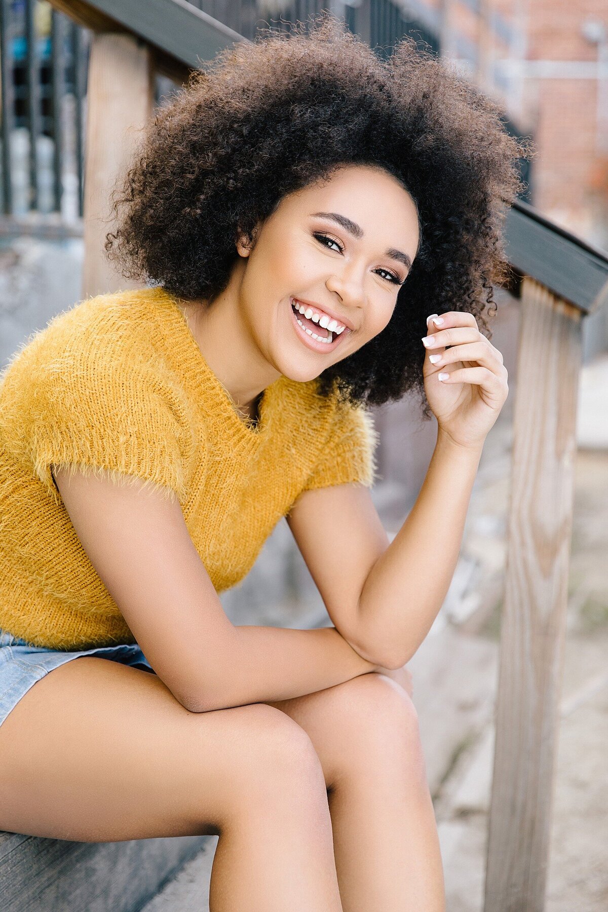 Fun and colorful senior portrait of curly-haired teen wearing mustard sweater and denim posing on urban steps in Los Angeles