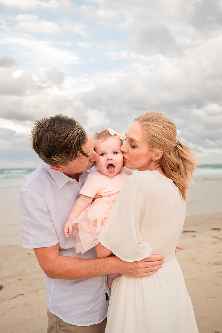 Candid adorable family photo of kid making face as parents kiss her on the Sandgate beach