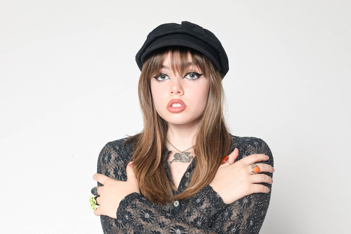 A person with long hair wears a black cap and a lace-patterned top, crossing their arms. They sport bold makeup and multiple rings, posing against a plain white background.