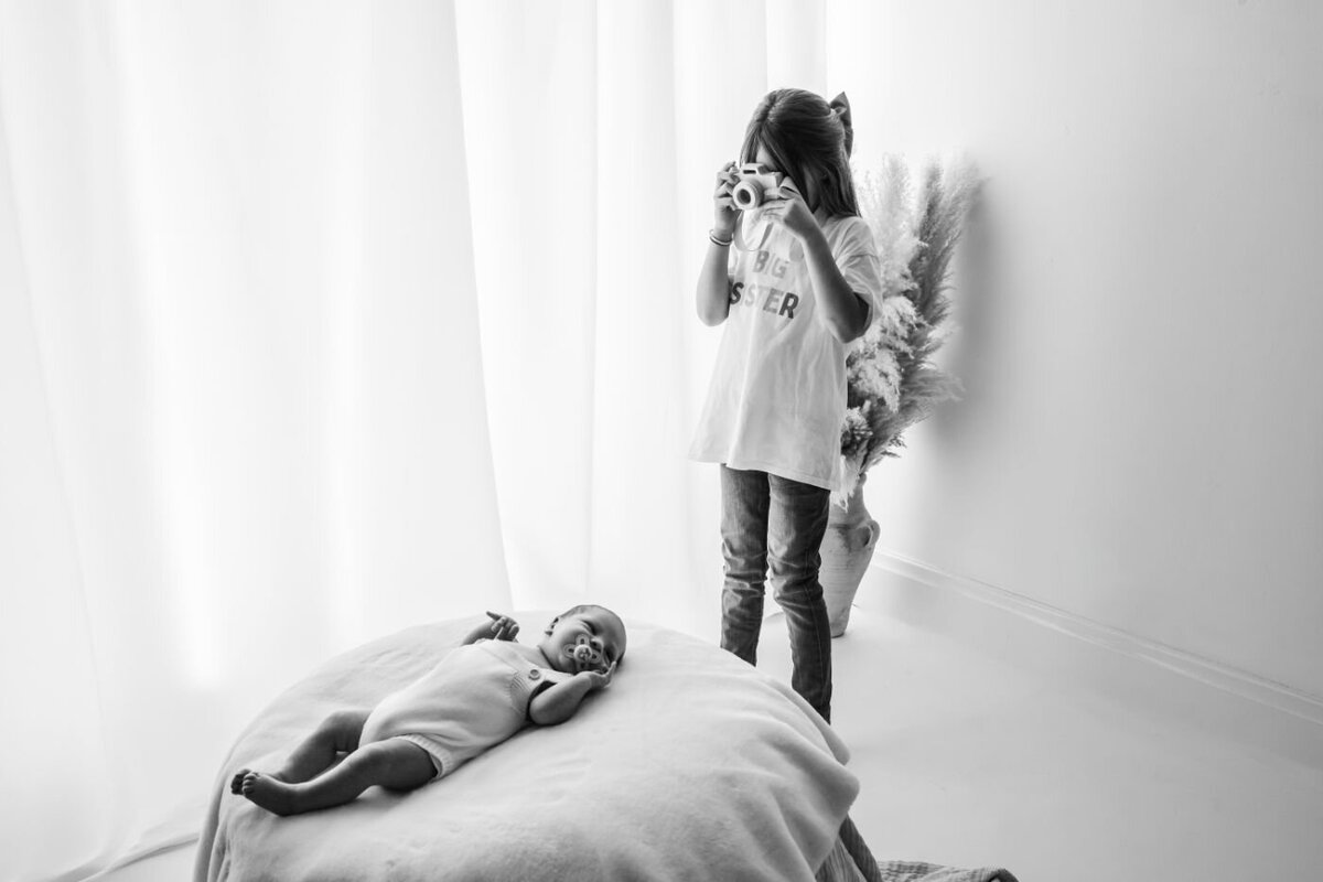 Monochrome shot of an older sister holding a camera and appearing to photograph her newborn sibling lying on a soft surface near a window in UK