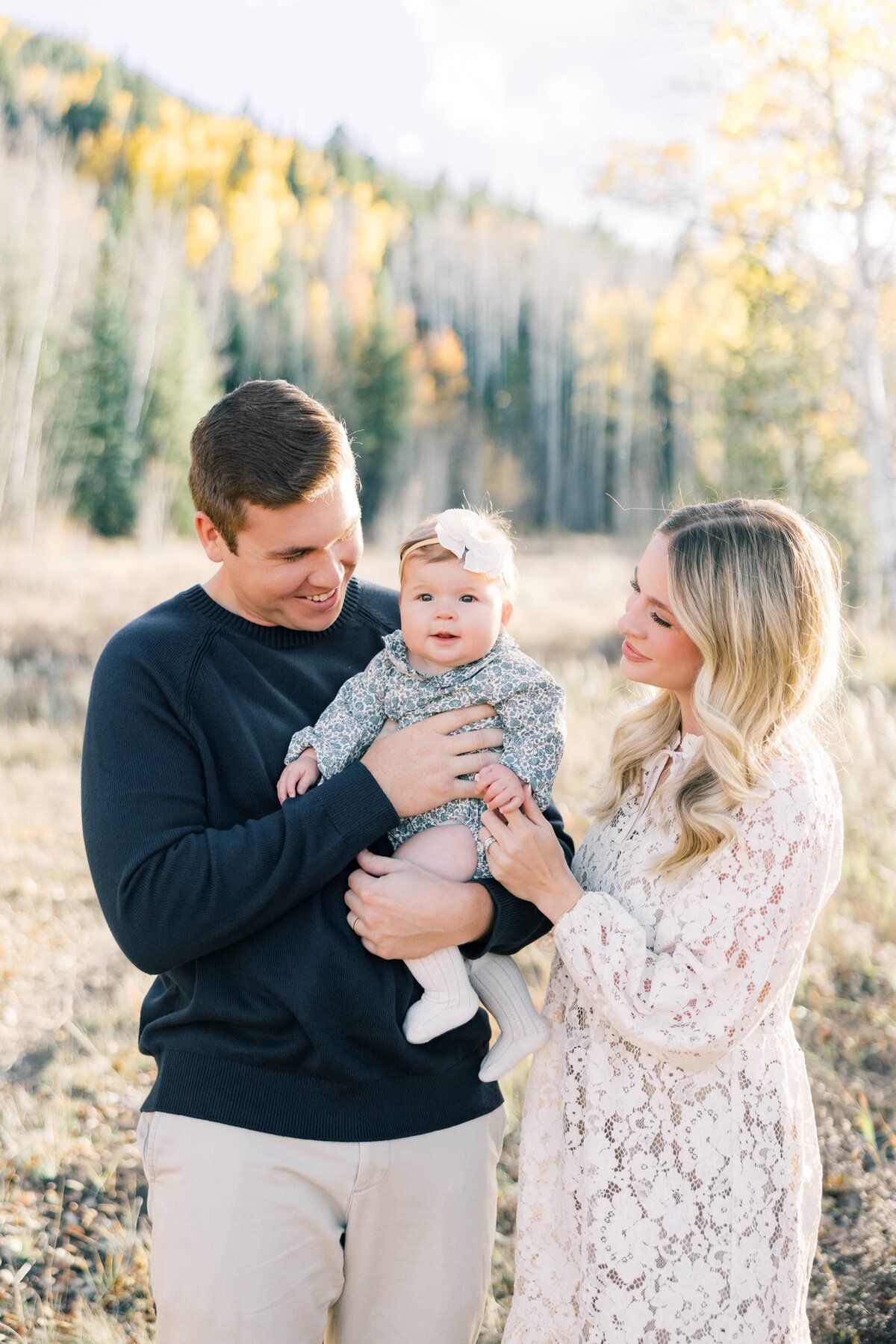 A young mom and dad are holding their 6 month old baby in their arms. There is a mountain scene with yellow aspens and mountains in the background. Taken by a local Breckenridge photographer. 
